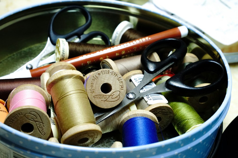 Photograph of a round tin container filled with spools of thread, small sewing scissors, and other sewing notions